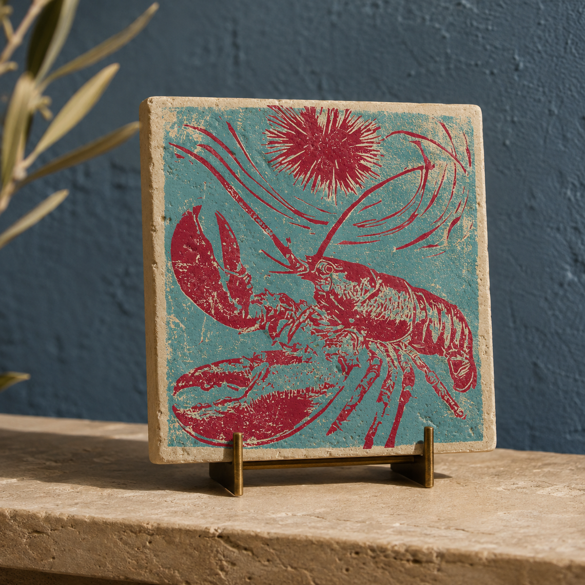 Quadratisches Travertin-Kunstwerk mit türkis-magentafarbenem Hummer, Seeigel und Wellenlinien auf einem Messingständer vor dunkelblauer Wand.
