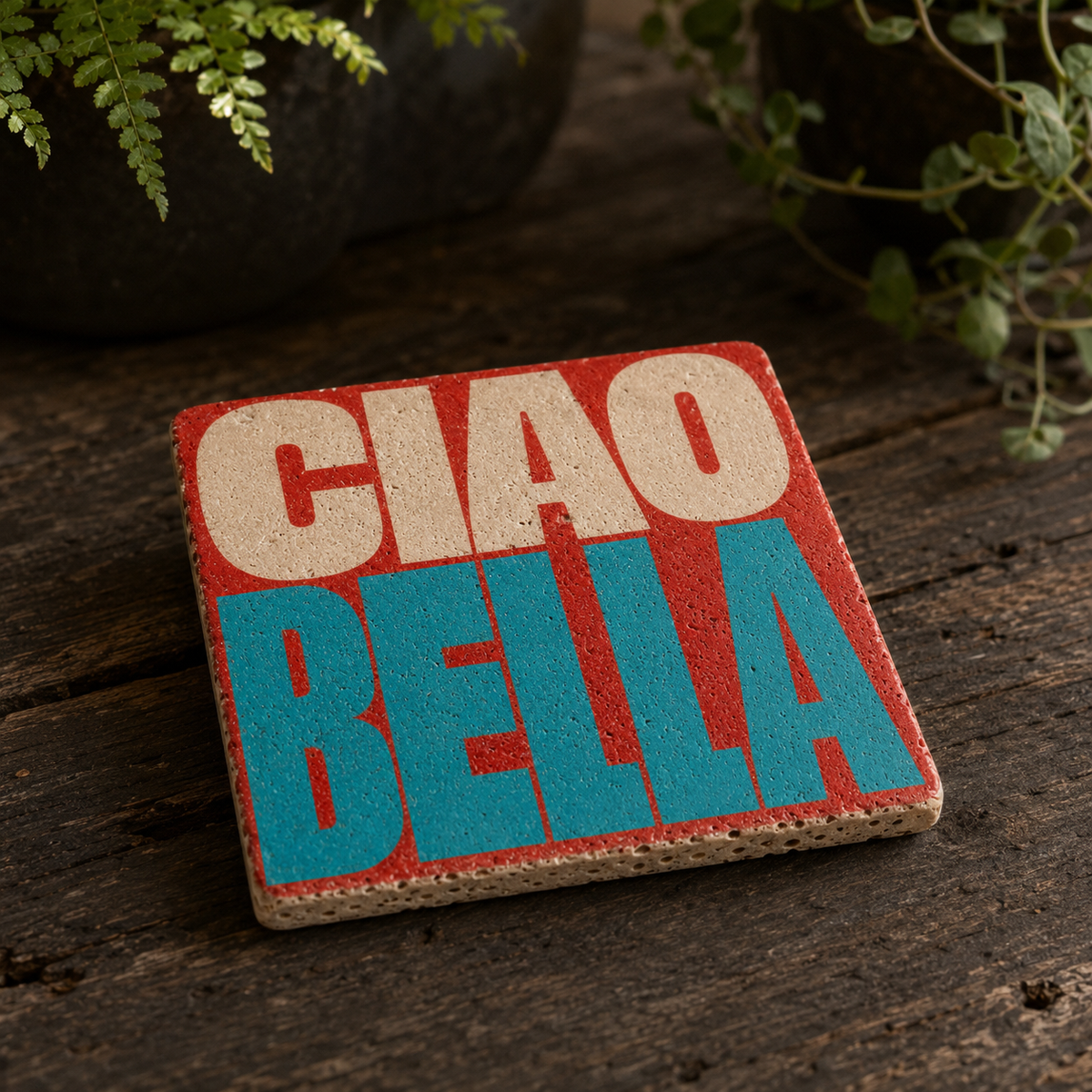 Ciao Bella Kunstwerk aus Travertin mit farbigem Schriftmotiv auf dunklem Holztisch mit grünen Pflanzen.