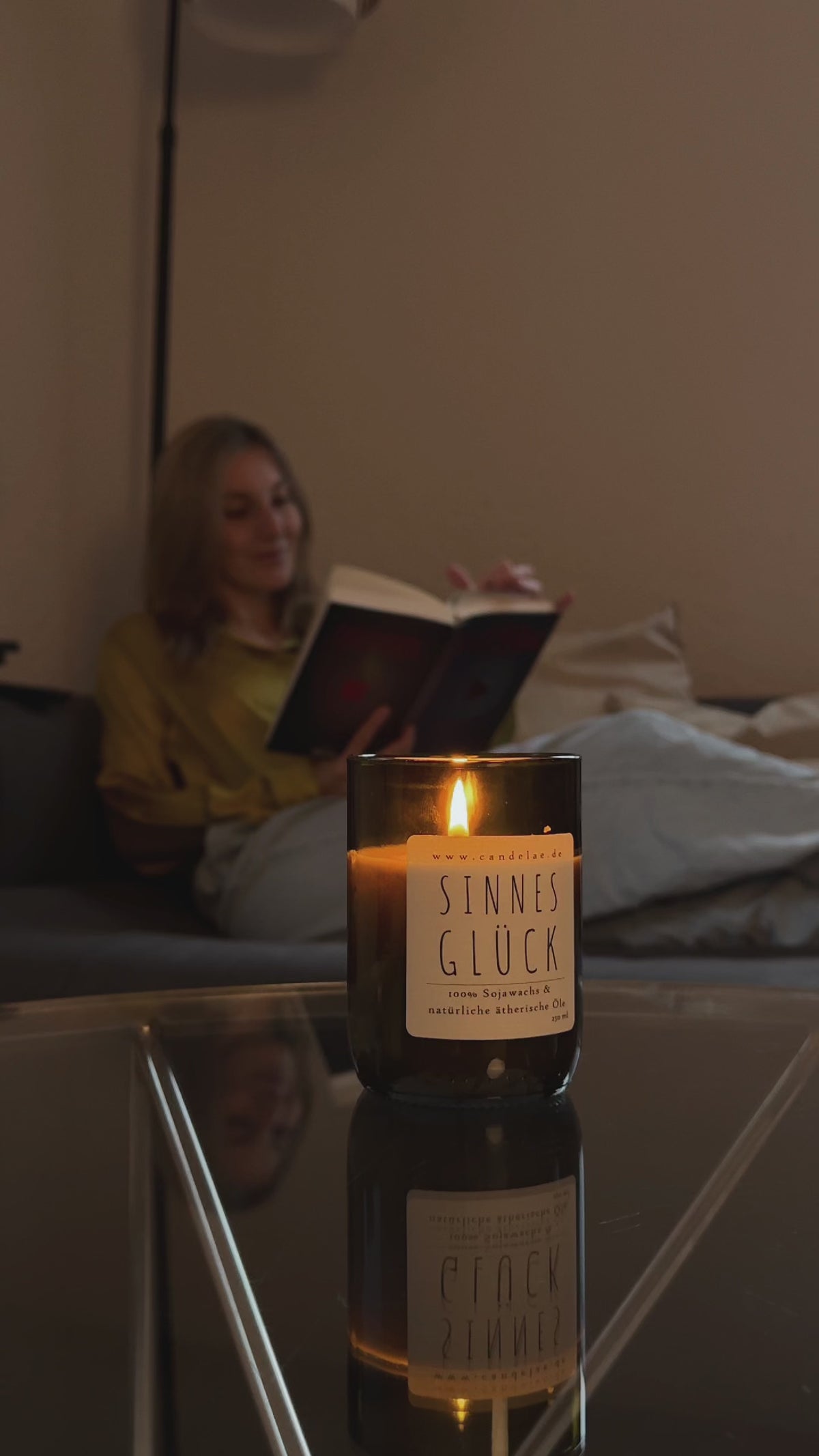 Sinnesglück von Candelae – Sojawachs-Duftkerze mit ätherischen Ölen im recycelten Weinflaschenglas brennt auf dem Couchtisch; im Hintergrund liest eine Frau.