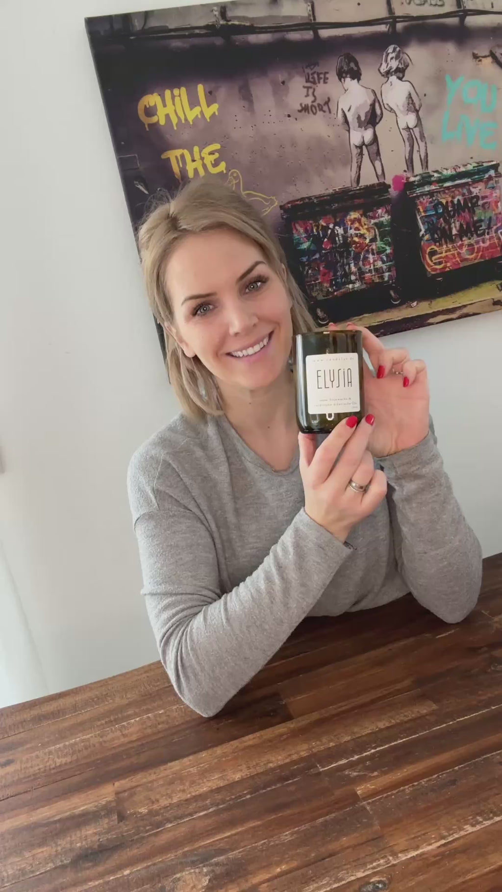 Kimberley präsentiert die Duftkerze Elysia in einem gemütlichen Ambiente. Sie hält die Kerze in der Hand, zeigt die Details des recycelten Weinglases und erzählt begeistert von den natürlichen Inhaltsstoffen und dem angenehmen Duft. Im Hintergrund sind eine schlichte, warme Dekoration und ein weiches Kerzenlicht zu sehen, die für eine einladende Atmosphäre sorgen.