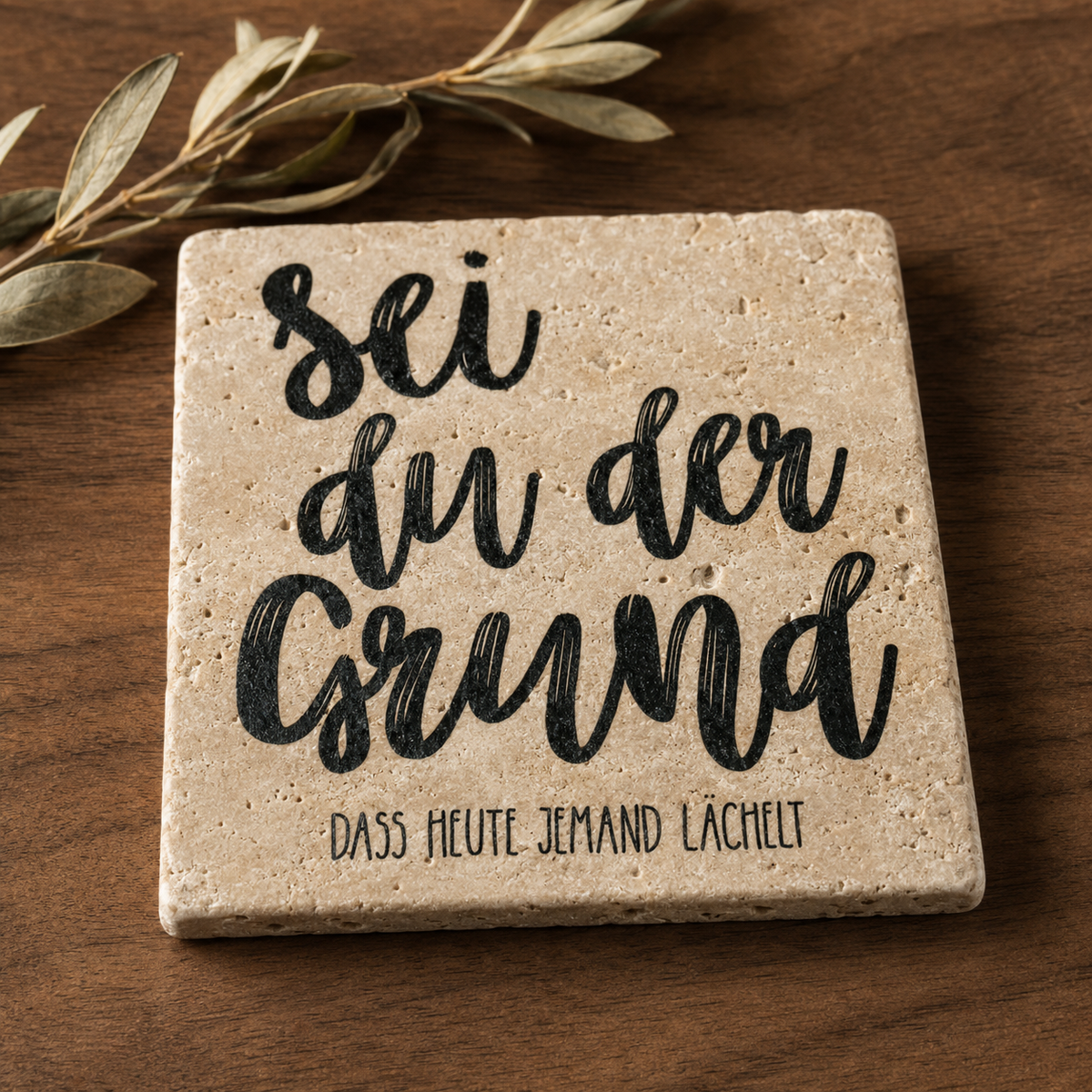 Kleines Kunstwerk aus Travertin mit dem schwarzen Spruch „Sei du der Grund, dass heute jemand lächelt“ auf einem Holztisch mit Olivenzweig.